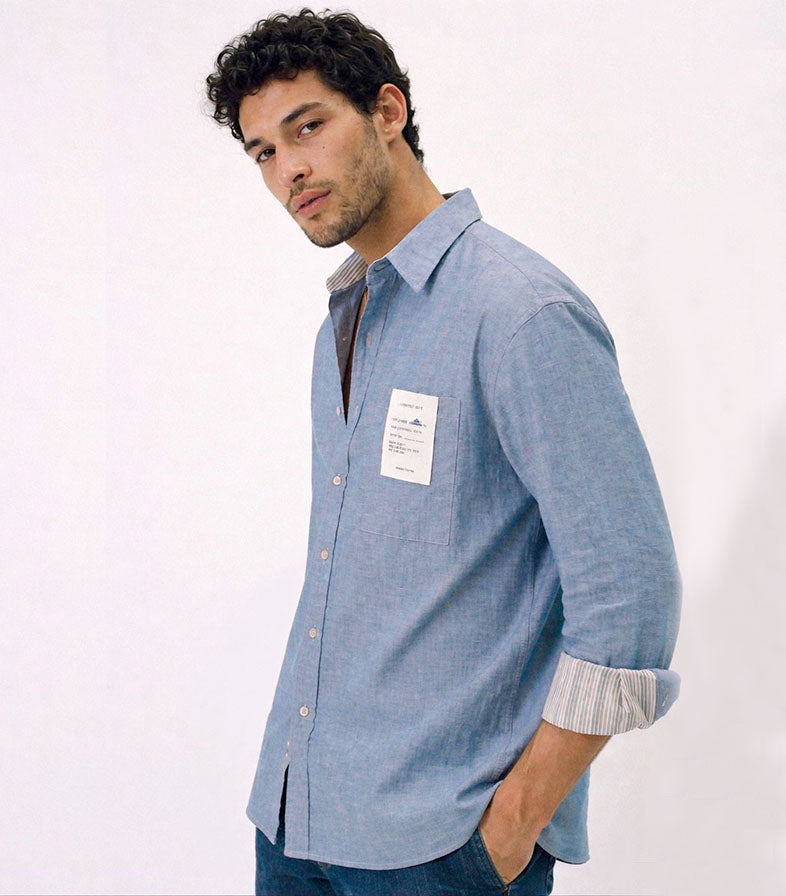 Camisa unisex de estilo  oversize con tejido algodón efecto denim y contraste en interior cuello y puños con un tejido de rayas, fit relajado estilo minimalista y moderno y urbano. estampado en la espalda y en el bolsillo delantero una etiqueta con número de serie exclusivo y personalizado